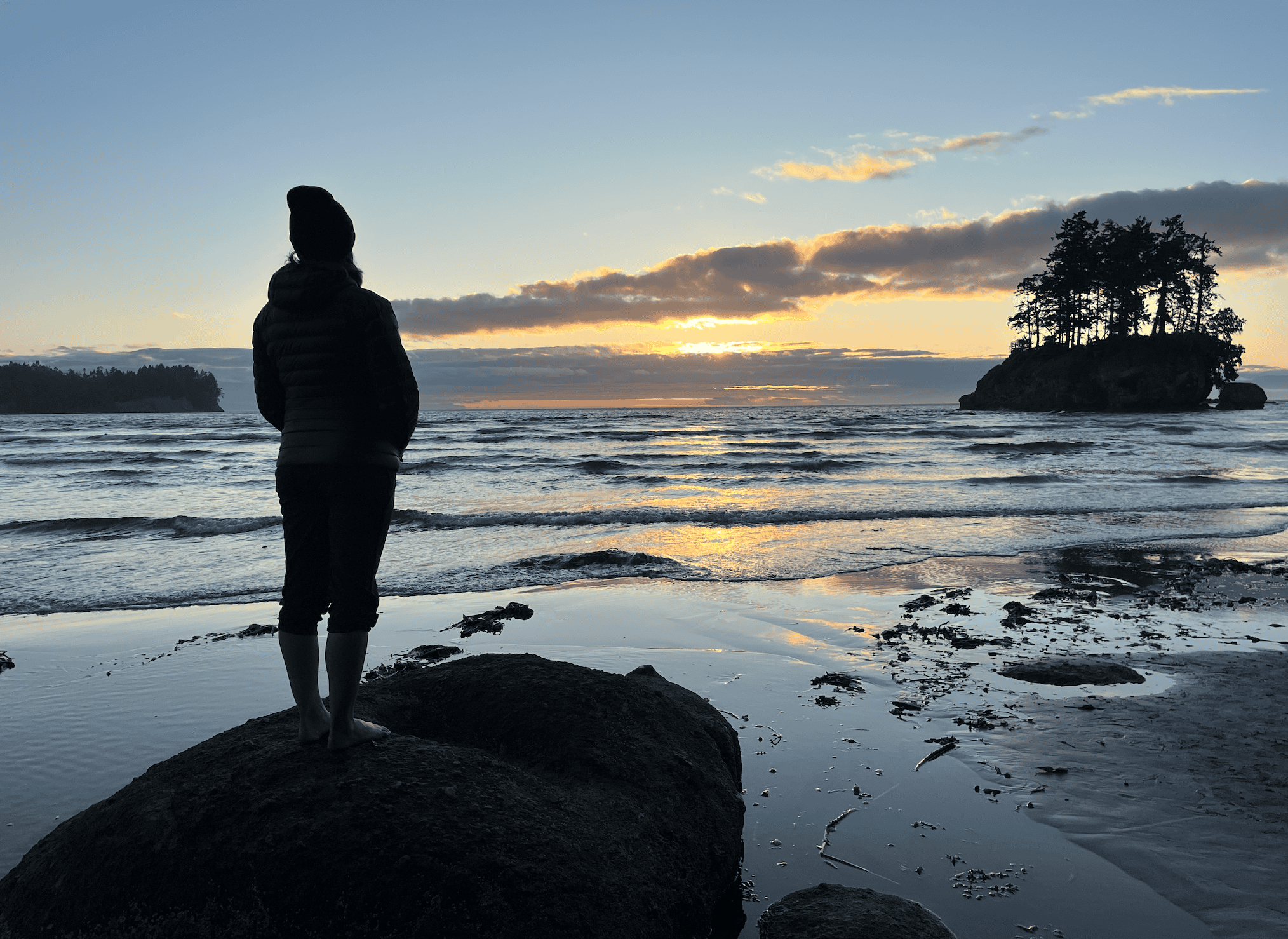 At sunset, a woman is standing on a rock overlooking the beautiful water in Port Angeles, Washington. This is a unique place to stay in Washington with charm, nature, and many outdoorsy activities.
